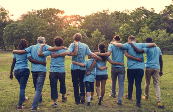 Eine bunt gemischte Gruppe von Personen mit blauen T-Shirts laufen sich gegenseitig umarmend vom Betrachter weg in Richtung eines Waldes in den Sonnenuntergang.